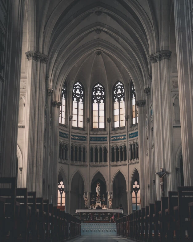 back of a majestic church with ornate architecture looking down the aisle toward the altar, view of a dark and empty church looking down the aisle between pews towards a beautiful altar with ornate church architecture in background