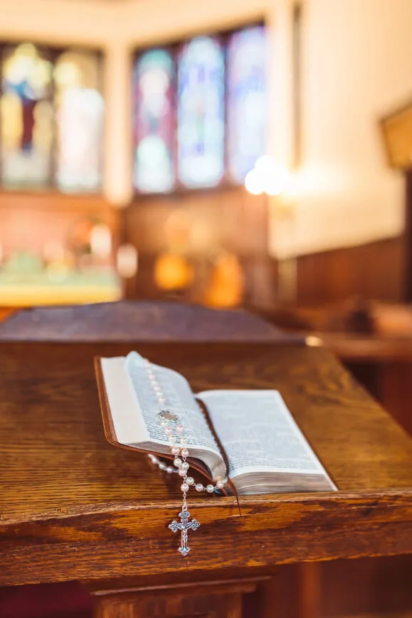 Rosary wrapped and entwined around an open Bible sitting on top of a kneeler, blurred background of the interior of a church, Rosary wrapped over open Bible that is sitting on kneeler with the interior of a church in the background